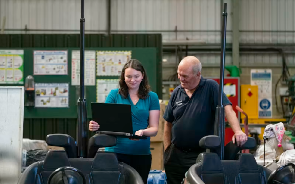 A woman with a laptop and an older man standing together in a workshop
